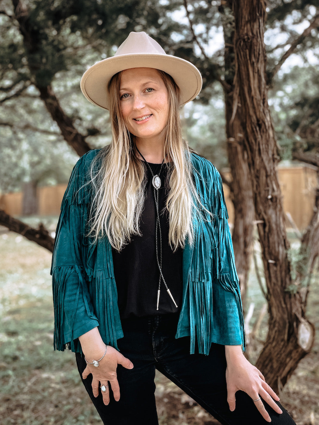 Woman smiling standing outside in front of trees wearing a cream colored cowboy hat, turquoise fringe jacket, black shirt and pants and a white oval Ivory Creek Variscite sterling silver bolo with black leather cord.