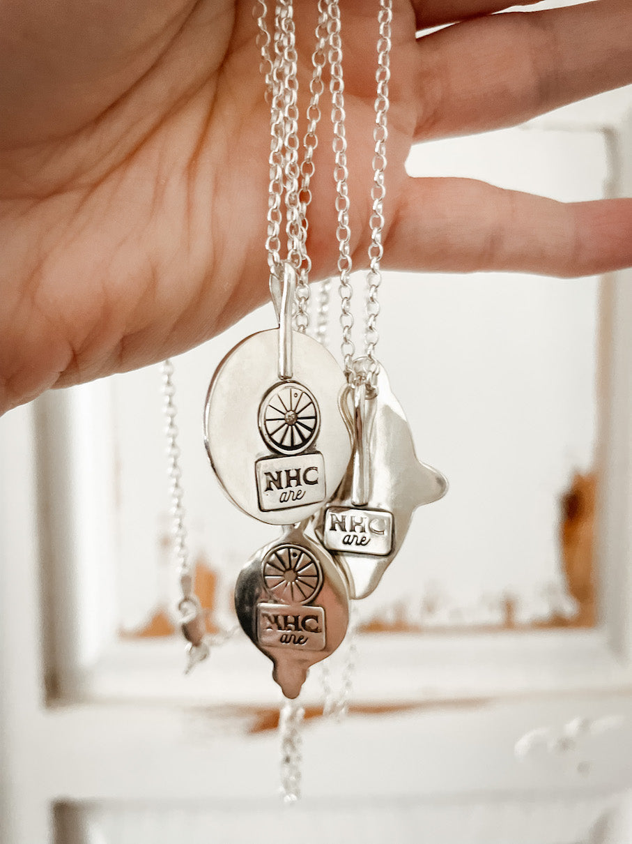 Hand holding three sterling silver necklaces hung from silver rollo chains against a distressed white and tan wood background.  The backs of the pendants are shown with the stamped Ninth House Collection maker's marks.