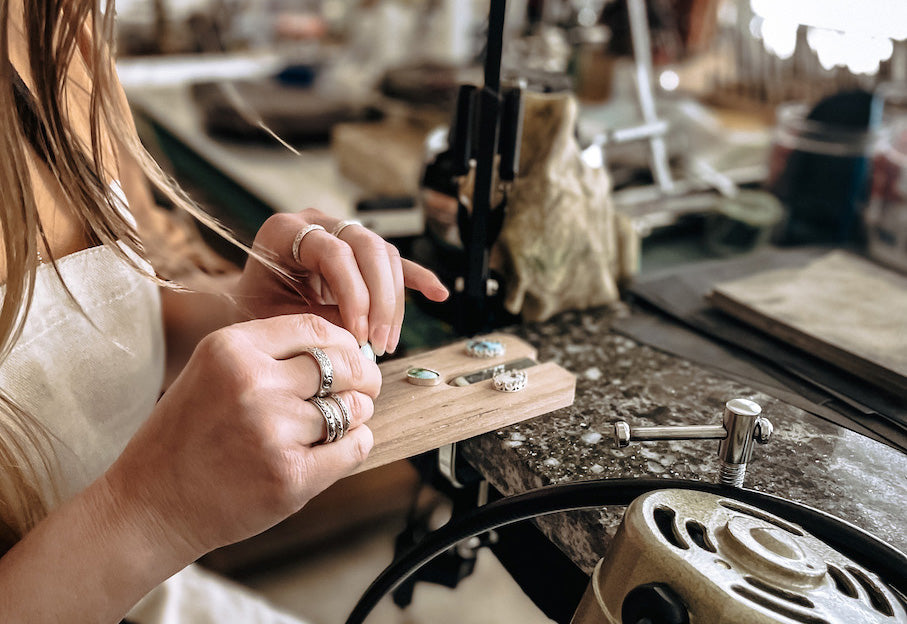 Woman in studio wearing silver rings and a white apron wrapping turquoise stones in silver bezels on a wooden bench pin attached to a marble slab.