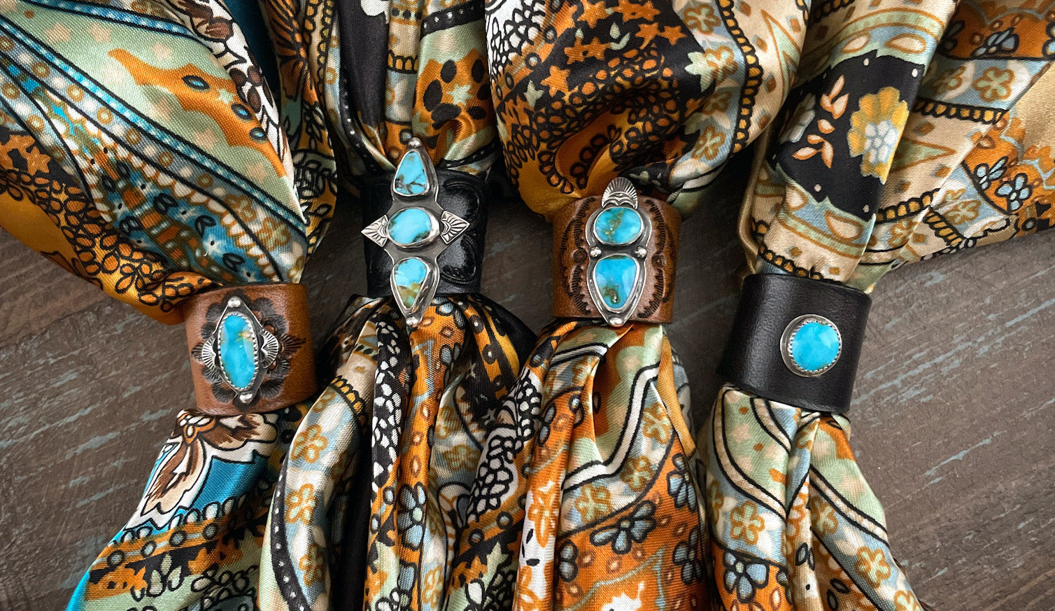 Four turquoise and silver bandana slides set on brown or black leather wrapped around brightly colored satin bandanas featuring orange, light green, blue, and black floral designs.
