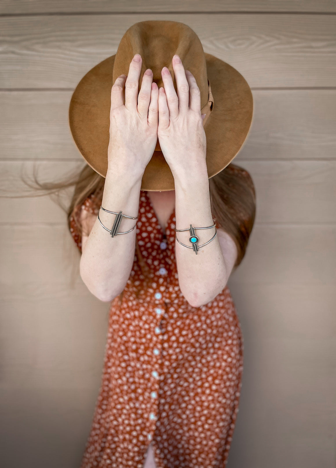 Woman standing in front of tan building wearing a rustic brown cowboy hat and brown short sleeve dress with white flowers on it. Her head is bent forward, her hair is blowing in the wind, and her hands are on top of her hat showing the large sterling silver cuff bracelets she is wearing on each arm. They are deco inspired with 3 vertical bars on each cuff. One cuff has a small round center turquoise stone in the middle.