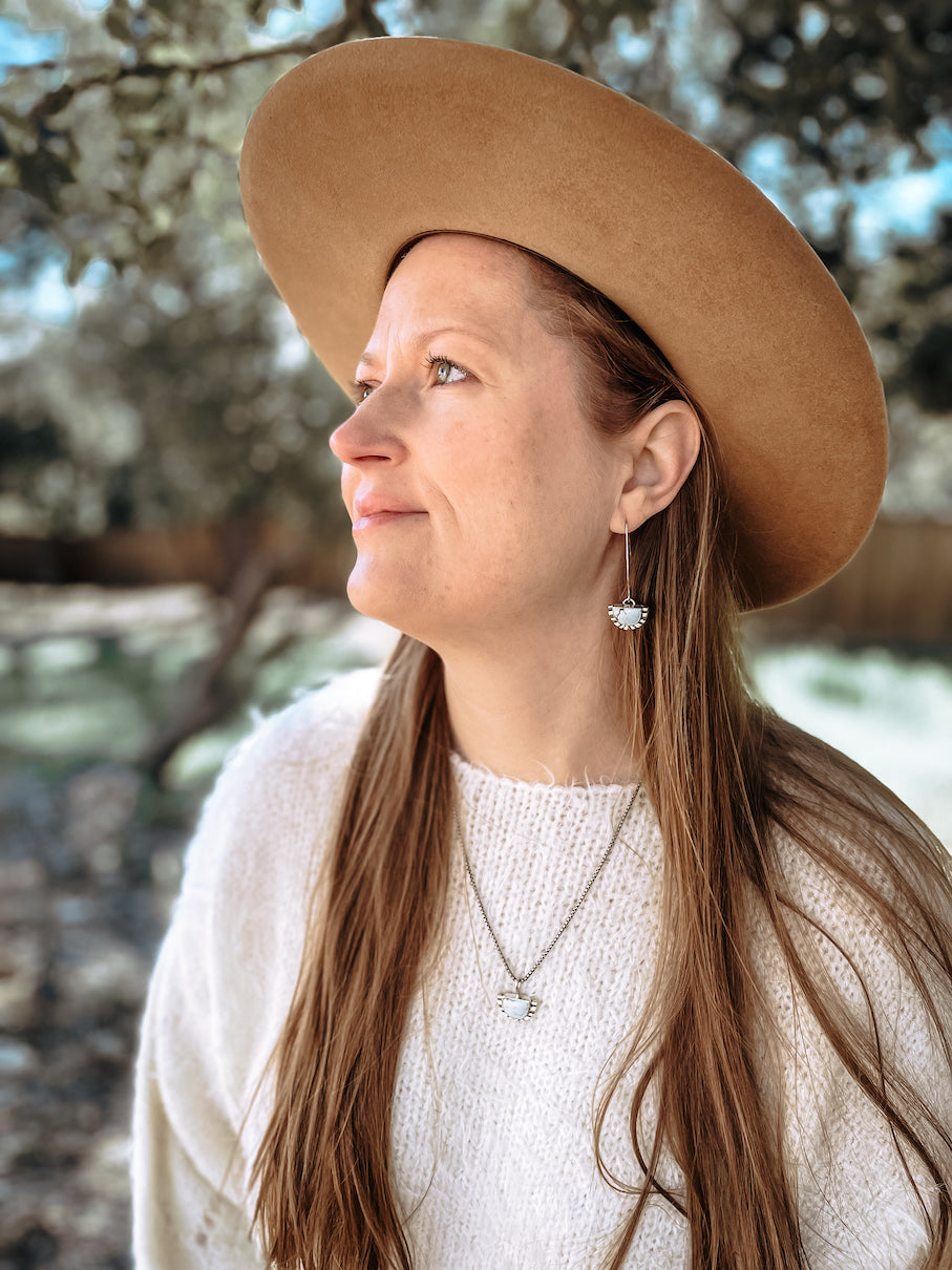 Woman with white sweater and brown western hat outdoors in natural light wearing half moon pale blue turquoise dangle earring in ear with matching sterling silver necklace.