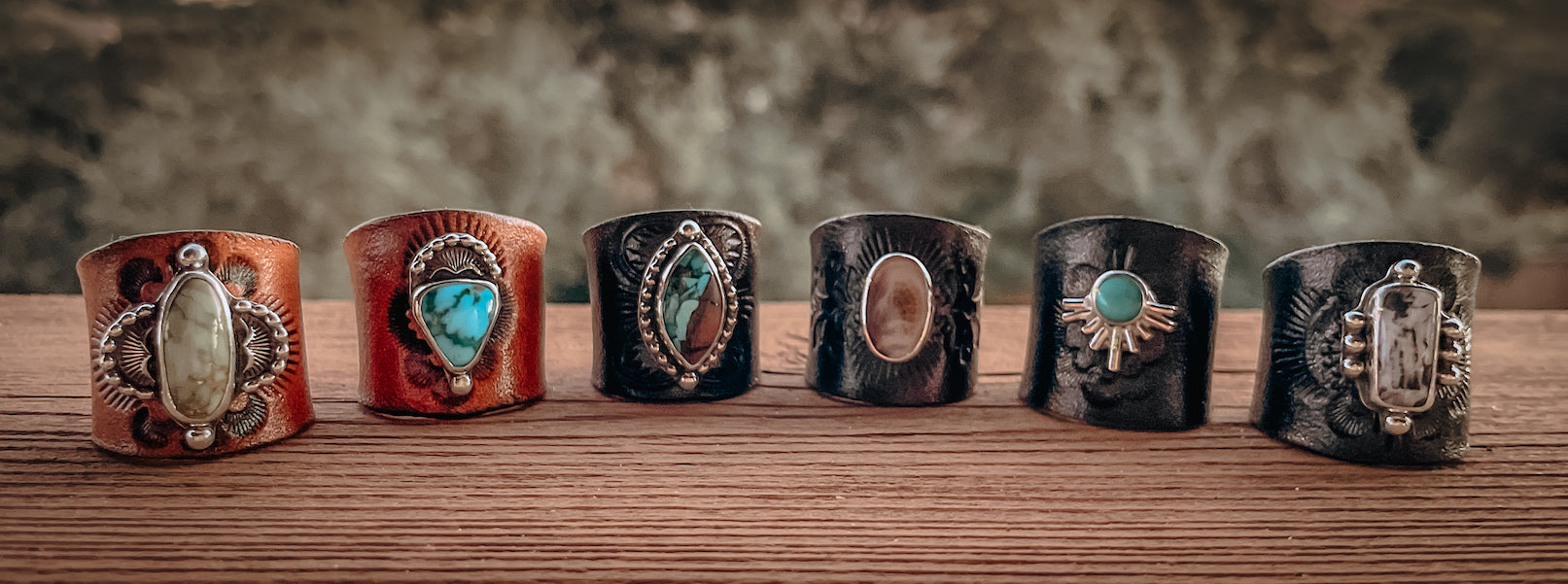 Six gemstone rag slides arranged on table outdoors featuring a variety of turquoise, white buffalo, and dendritic stones set in sterling silver attached to stamped leather slides in tan, brown, and black colors.