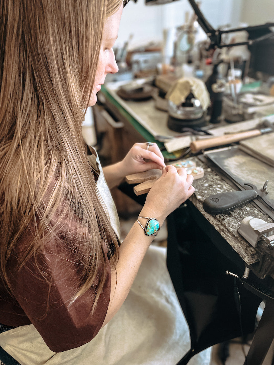 Woman sitting in studio wearing a large teardrop turquoise cuff bracelet and a white apron wrapping stones in silver bezels on a wooden bench pin attached to a marble slab.