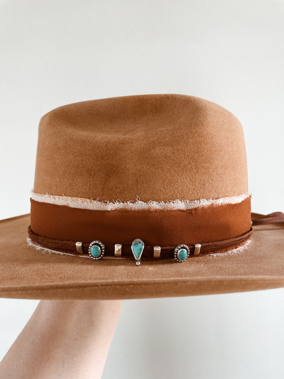 Small turquoise hat slides on hat featuring center teardrop stone slide with two silver plain band accent slides on each side and round stone slides alternating with plain band slides on the ends on a thin brown leather hat band layered on top of a fabric hatband.