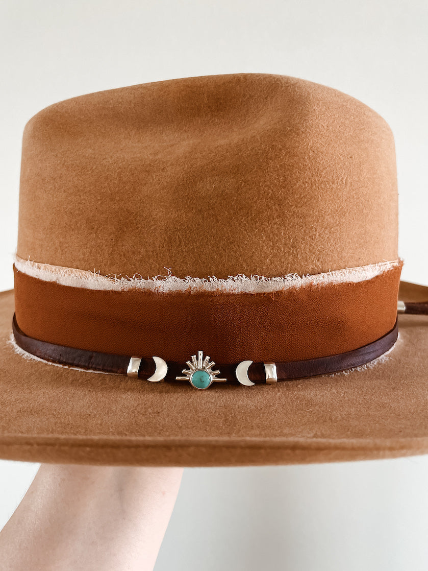 Small turquoise hat slides on hat featuring center round turquoise stone starburst slide with silver crescent moon accent slides on each side and a plain band accent slide on each end on a thin brown leather hat band layered on top of a fabric hatband.