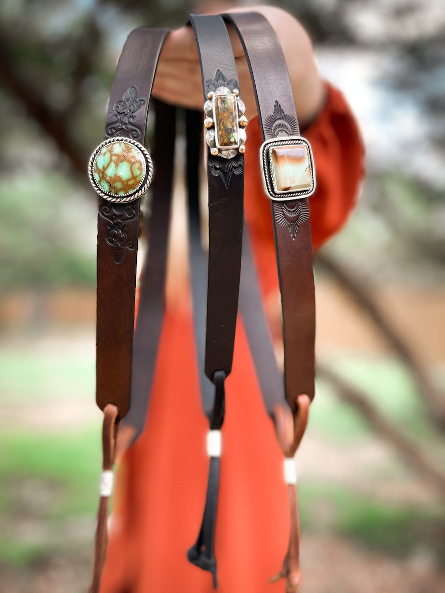 Woman in orange dress holding three hatbands featuring round, rectangle, and square turquoise in sterling silver settings on dark leather bands with stamped designs.