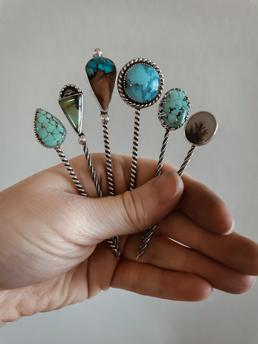 Six stick hat pins held in hand featuring turquoise, wood & turquoise inlay, and dendritic agate stones.