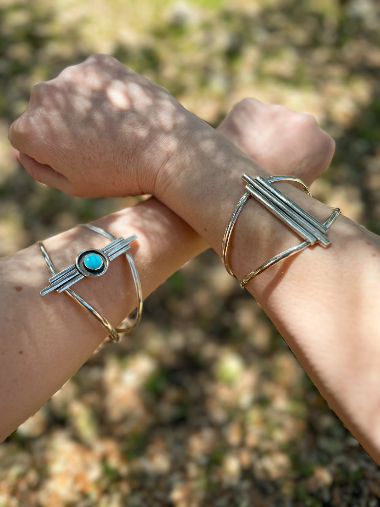 Two Art Deco inspired sterling silver cuff bracelets worn on crossed wrists outdoors in natural light. One features a small round turquoise stone in the center.