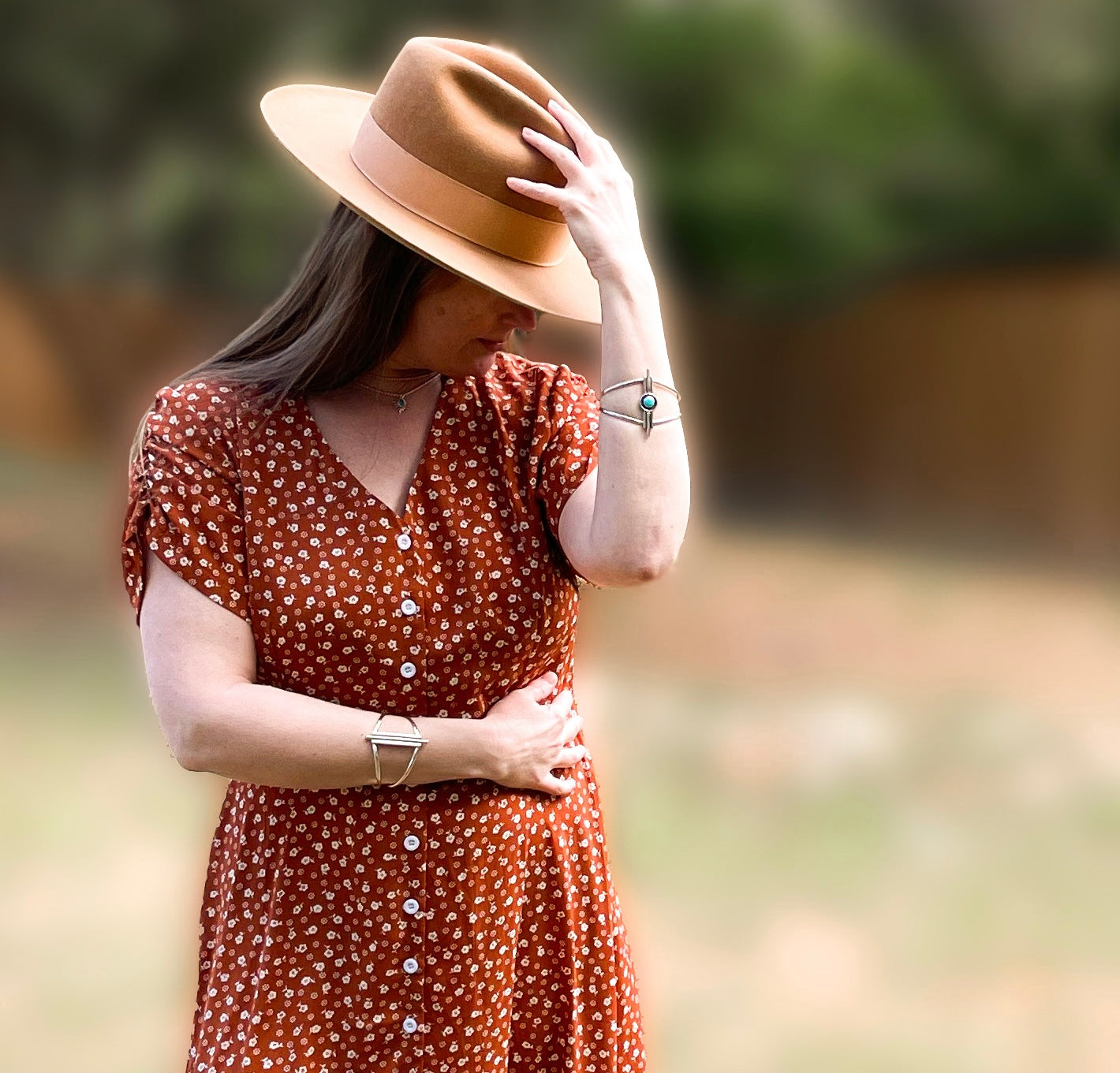 Woman wearing a brown dress with white flowers wearing Art Deco inspired sterling silver cuff bracelets on each wrist outdoors. She is wearing a brown western style hat with her head bent down to one side. One hand is touching the top of the hat and her other hand is across her waist to show the cuff designs.