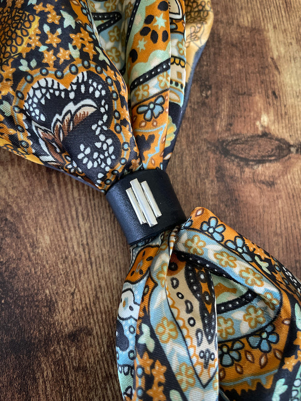 Art Deco inspired silver and black leather wild rag scarf slide on a colorful orange, black, pale green and blue silk scarf laying on a wood table.