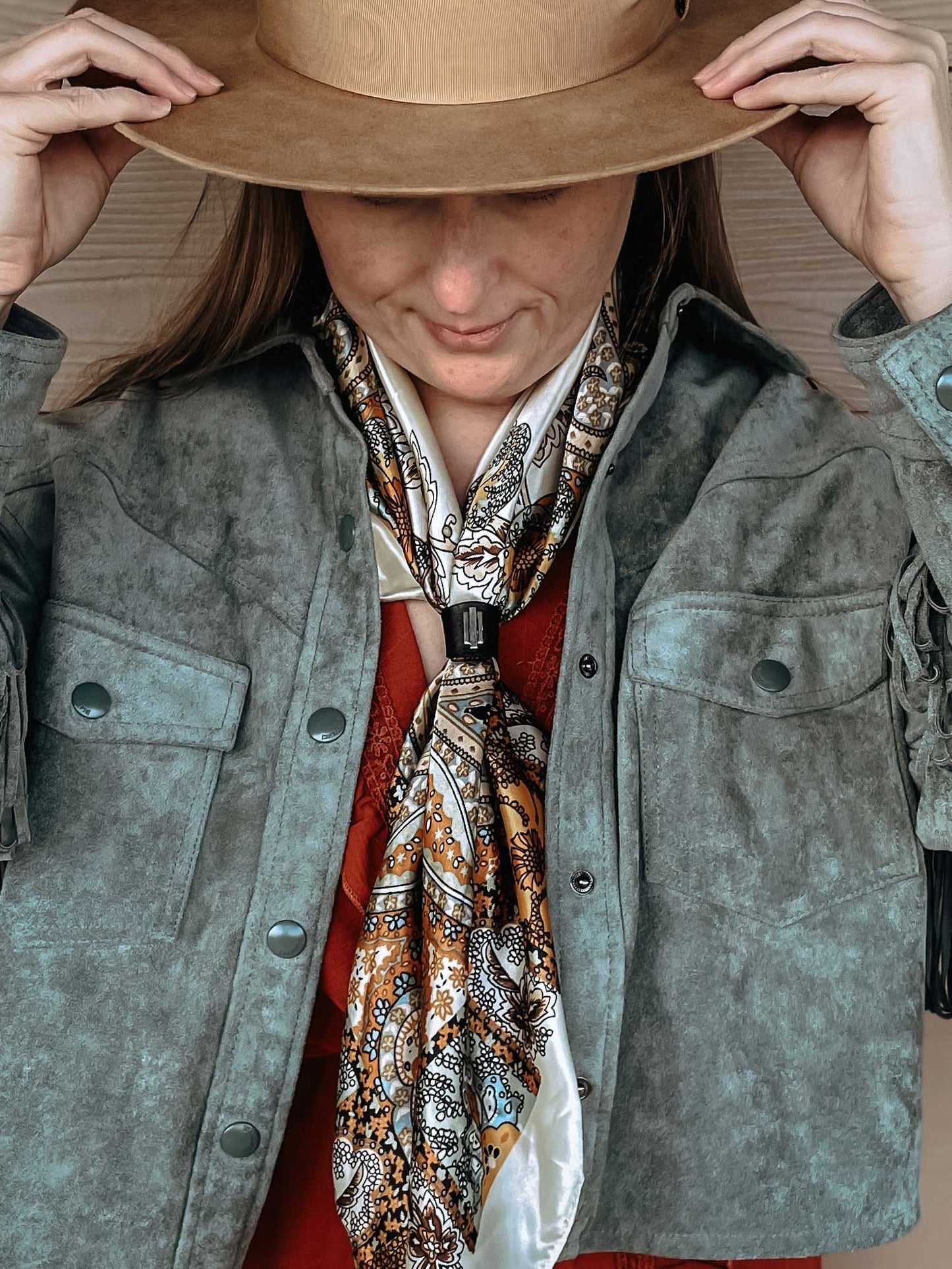 Woman wearing Art Deco inspired silver and black leather wild rag scarf slide on a cream colored orange, black, and pale blue silk scarf. She is wearing a light blue fringe jacket and brown cowboy hat.