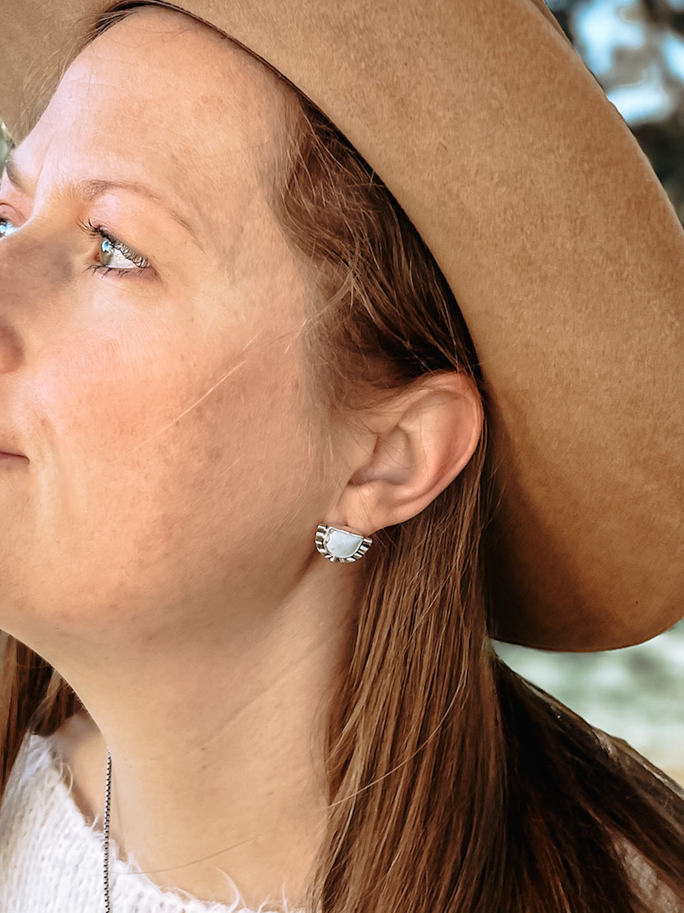 Woman with white sweater and brown western hat outdoors in natural light wearing half moon pale blue turquoise sterling silver stud earring in ear.