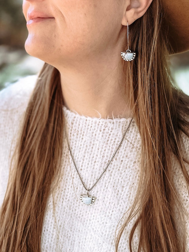 Woman with white sweater outdoors in natural light wearing half moon pale blue turquoise dangle earring in ear with matching necklace.