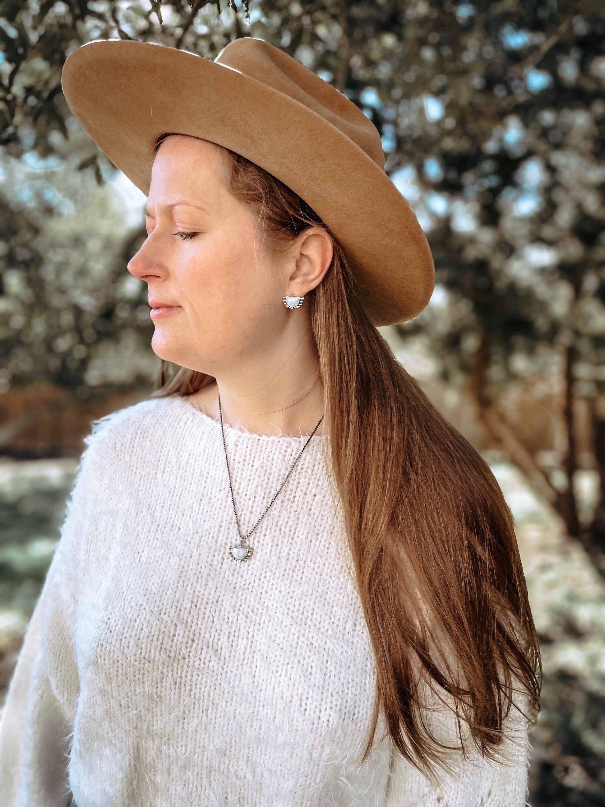 Woman with white sweater and brown western hat outdoors in natural light wearing half moon pale blue turquoise stud earring in ear with matching sterling silver necklace.
