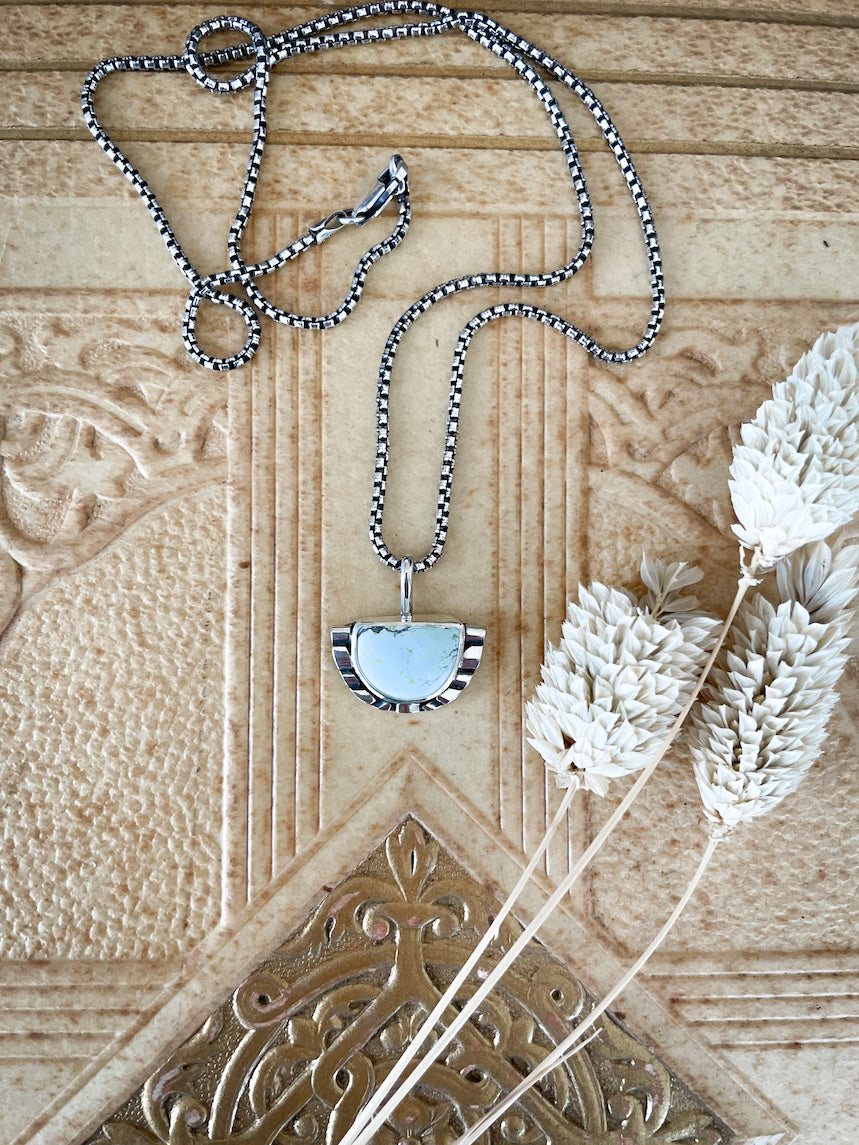 Half moon pale blue turquoise pendant necklace featuring hand-shaped silver moonbeams accenting the curves of the stones on a rounded box chain laying on brown Art Deco light brown book cover with cream colored flowers placed on it.