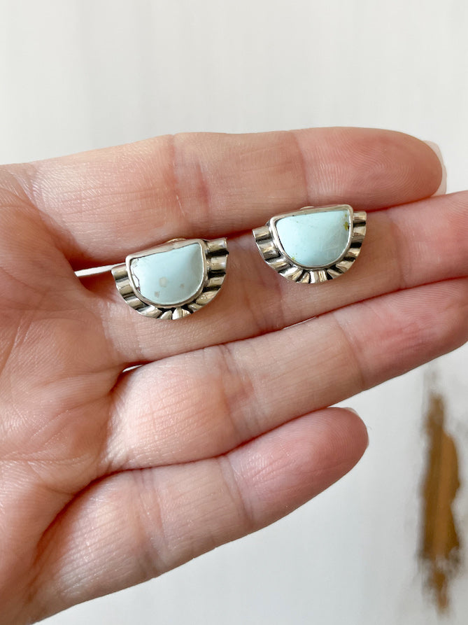 Pair of half moon pale blue turquoise sterling silver stud earrings held in hand against white wood background.