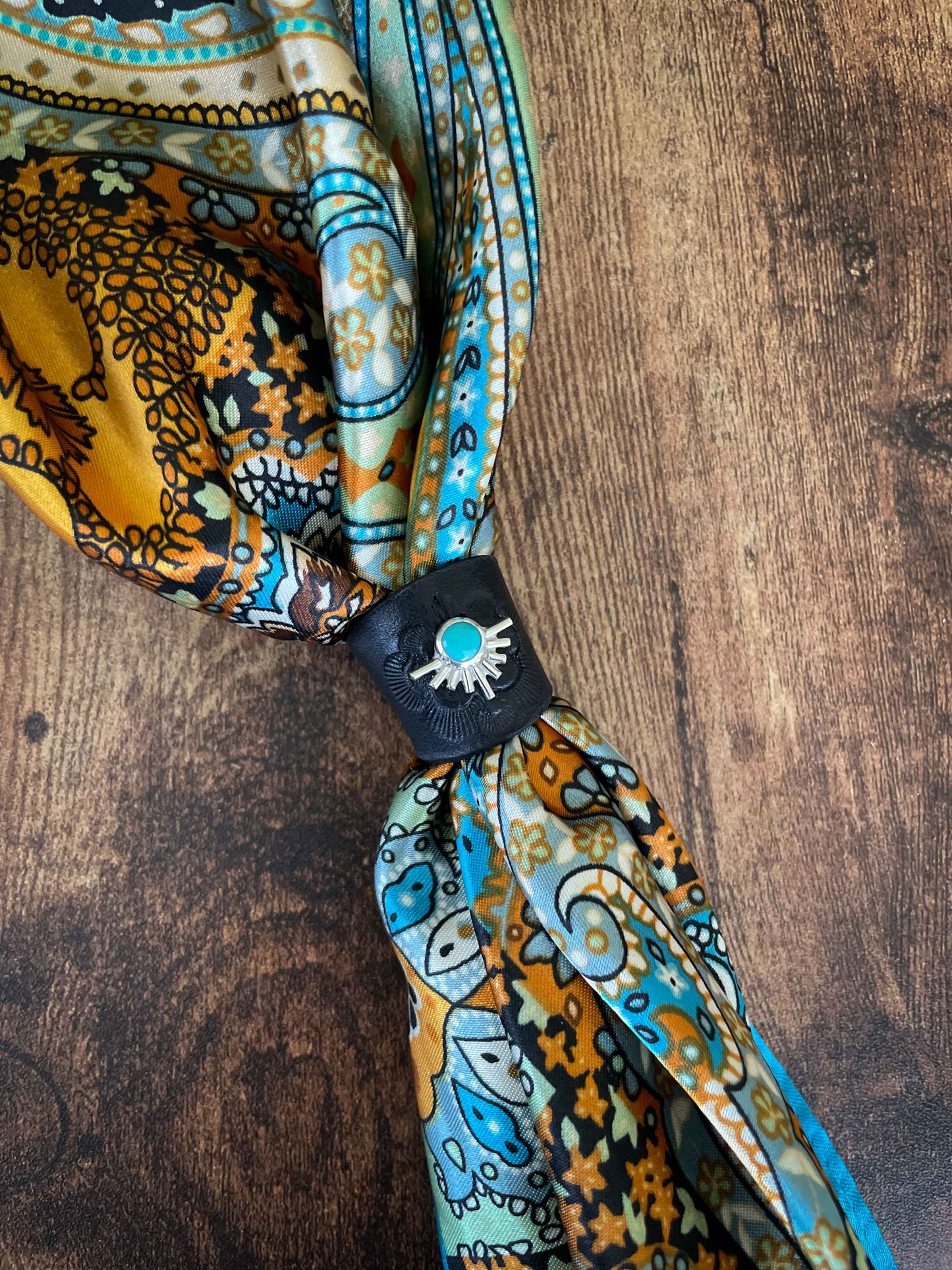 Sterling silver, turquoise, and black leather wild rag scarf slide on a colorful orange, black, pale green and blue silk scarf laying on a wood table.