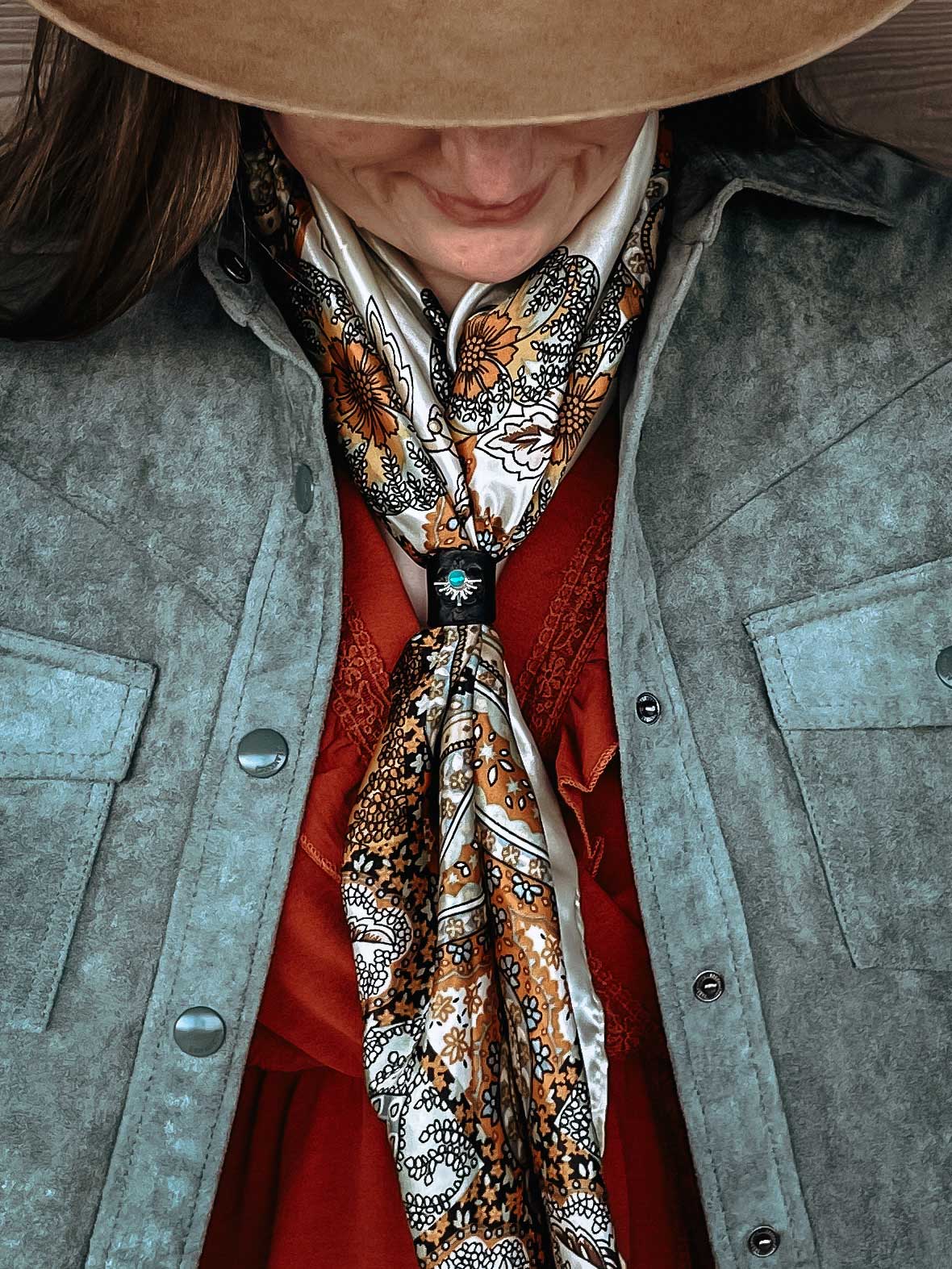 Woman wearing a sterling silver, turquoise, and black leather wild rag scarf slide on a cream colored orange, black, and pale blue silk scarf. She is wearing a light blue fringe jacket and brown cowboy hat.